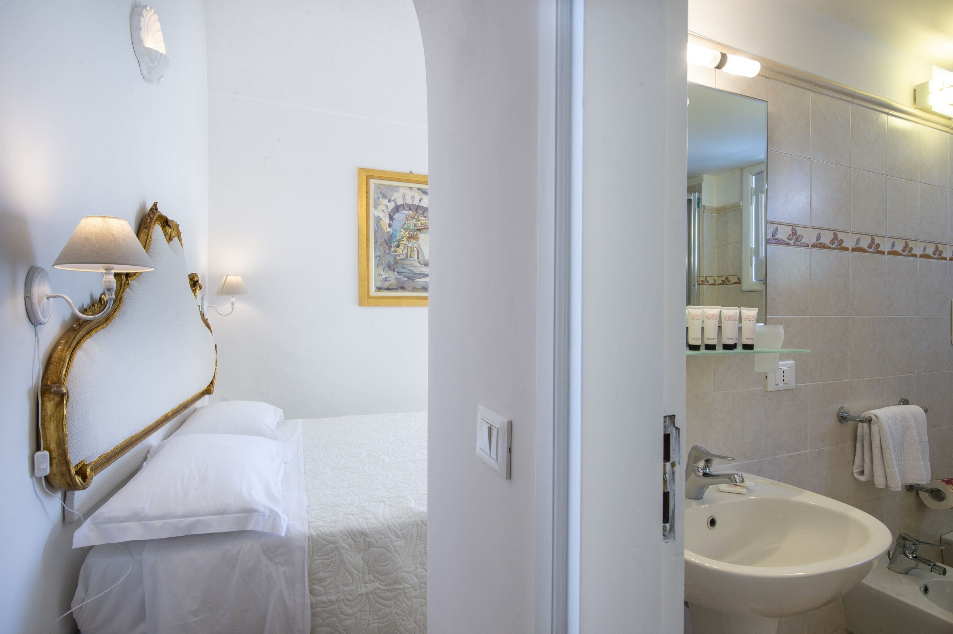 A hotel room with a bed, a wall-mounted lamp, a framed painting, and a bathroom with a sink, mirror, and towel rack.