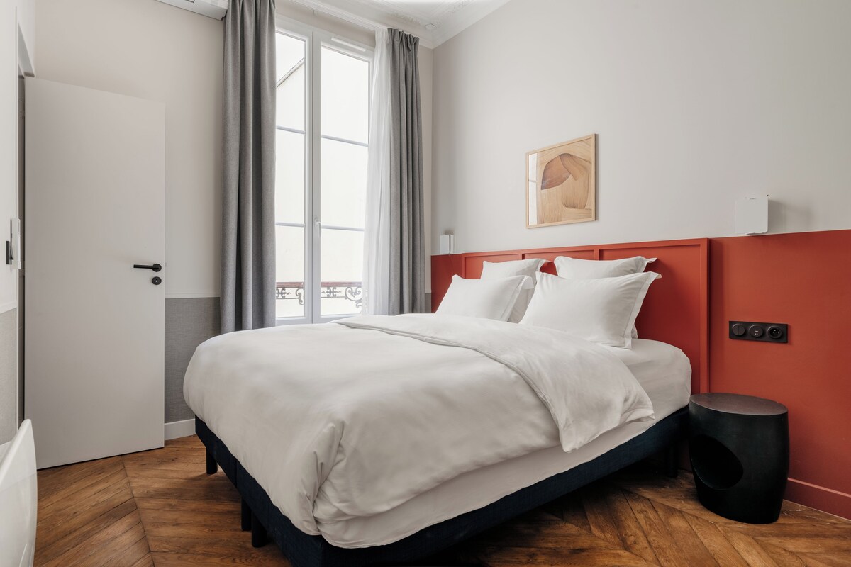 Une chambre d’hôtel moderne avec un grand lit, un mur d’accent rouge, une fenêtre avec des rideaux et une petite table d’appoint.