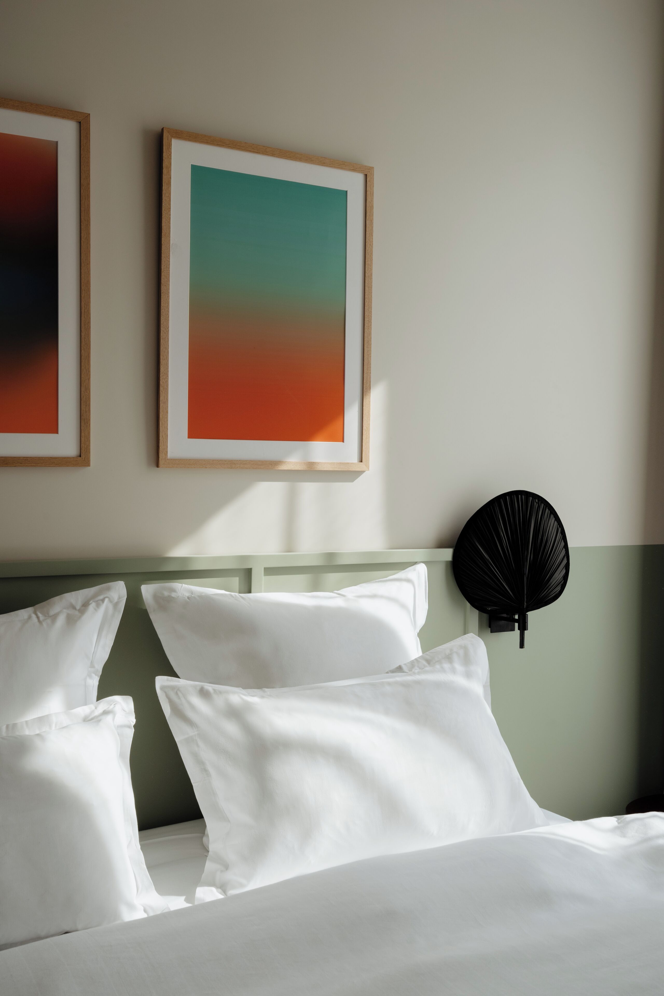 A hotel room with a bed, pillows, a wall-mounted fan, and two framed artworks.