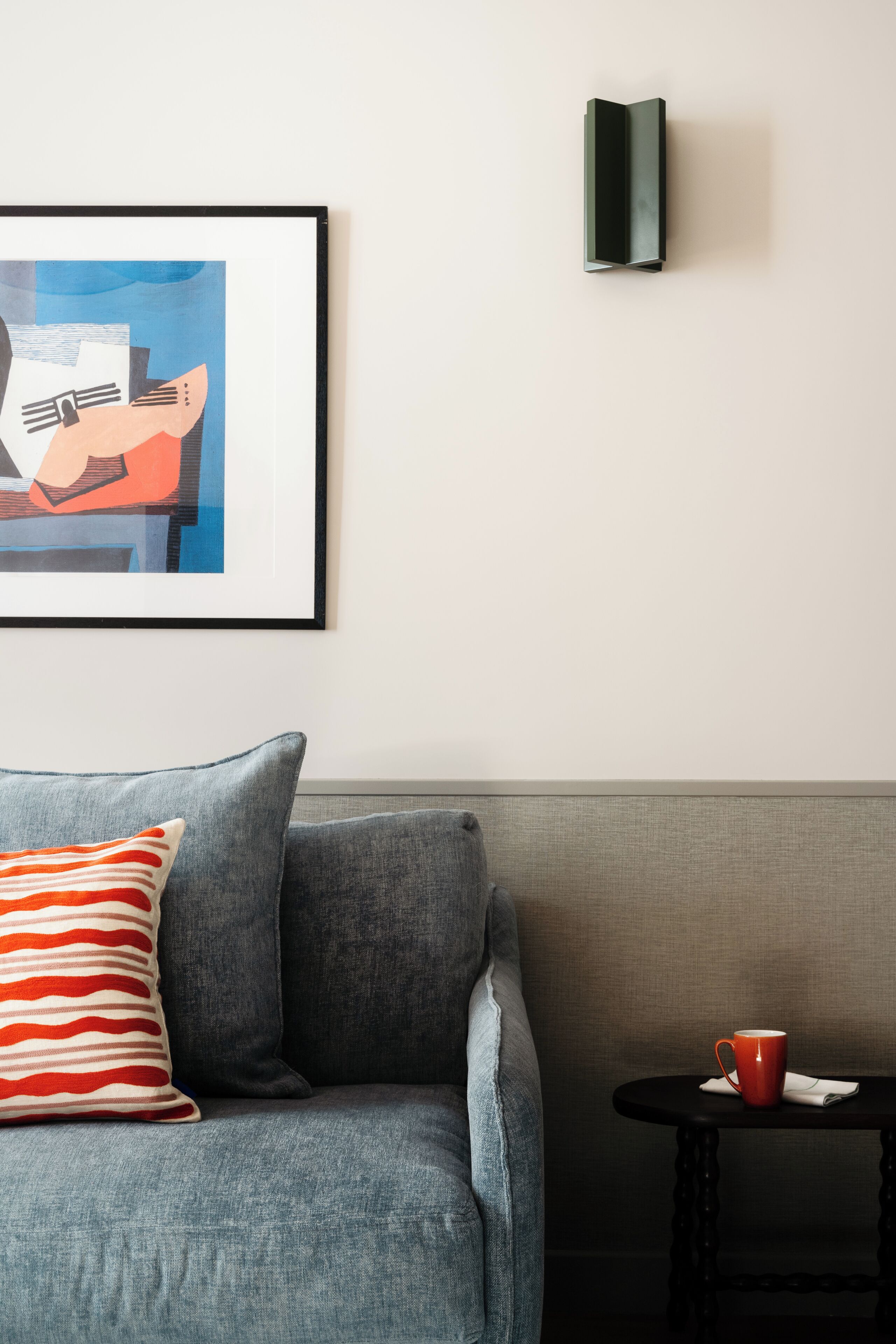 Une pièce avec un canapé gris, une œuvre d’art encadrée, une petite table sur laquelle se trouve une tasse rouge et un luminaire fixé au mur.