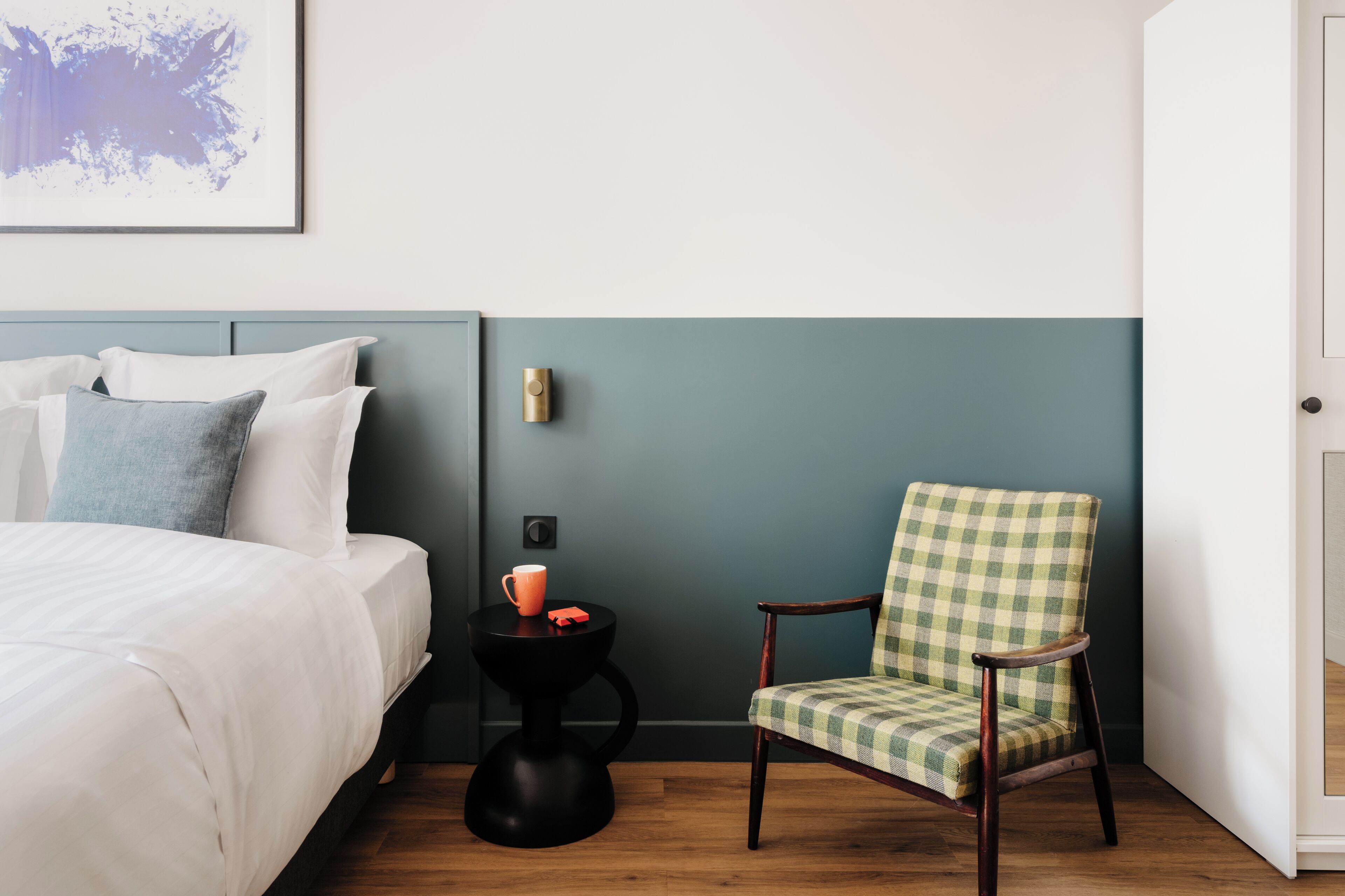 A hotel room with a bed, a chair, a small table, and a wall-mounted light fixture.
