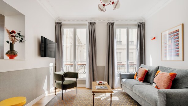 Una sala de estar moderna con un sofá gris, una mesa de centro de madera, un taburete amarillo y una alfombra estampada en gris.