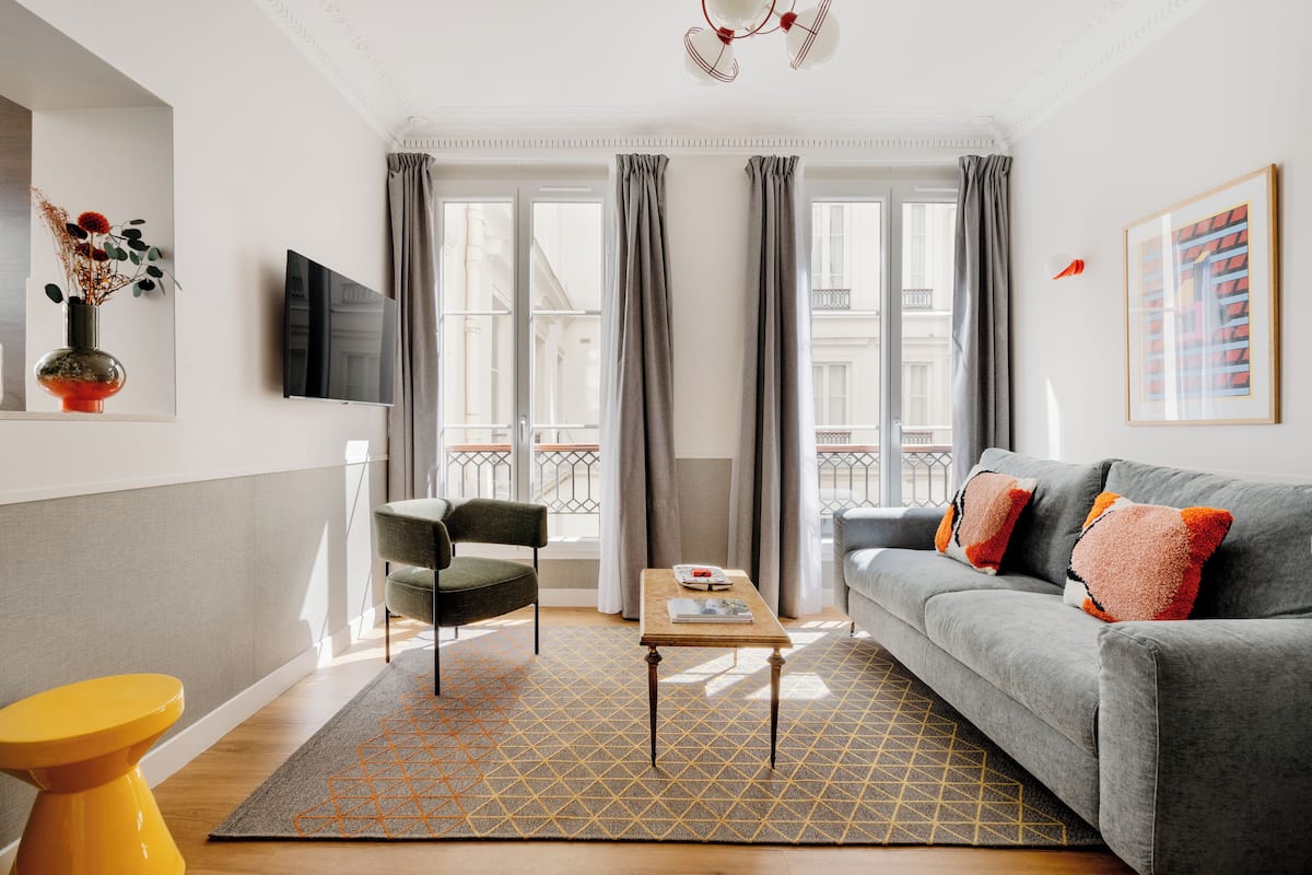 Una sala de estar moderna con un sofá gris, una mesa de centro de madera, un taburete amarillo y una alfombra estampada en gris.
