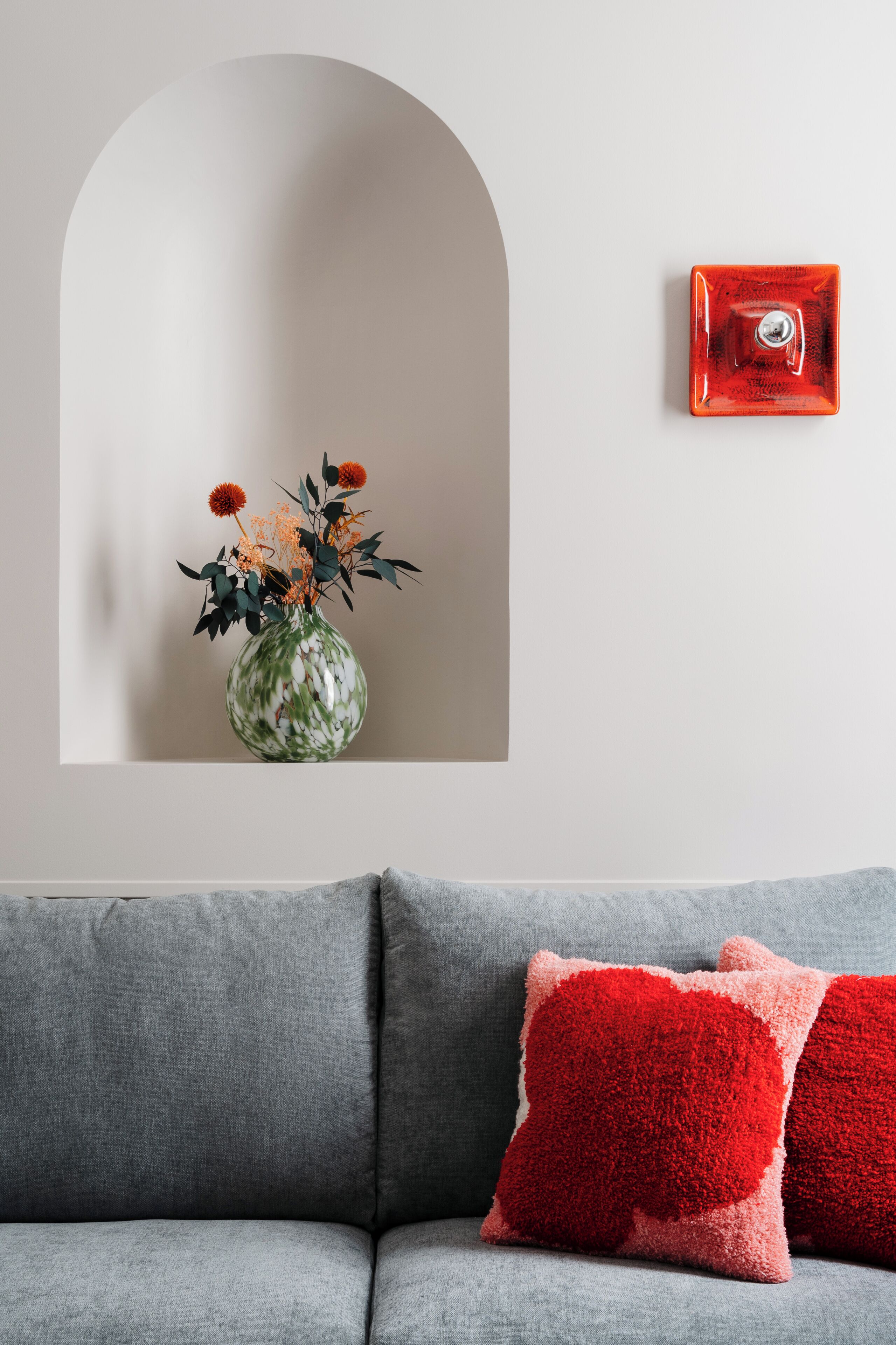 Ein graues Sofa mit roten und grauen Kissen, ein rotes, rechteckiges Objekt an der Wand und eine grüne, weiße Vase mit Blumen, ebenfalls an der Wand befestigt.