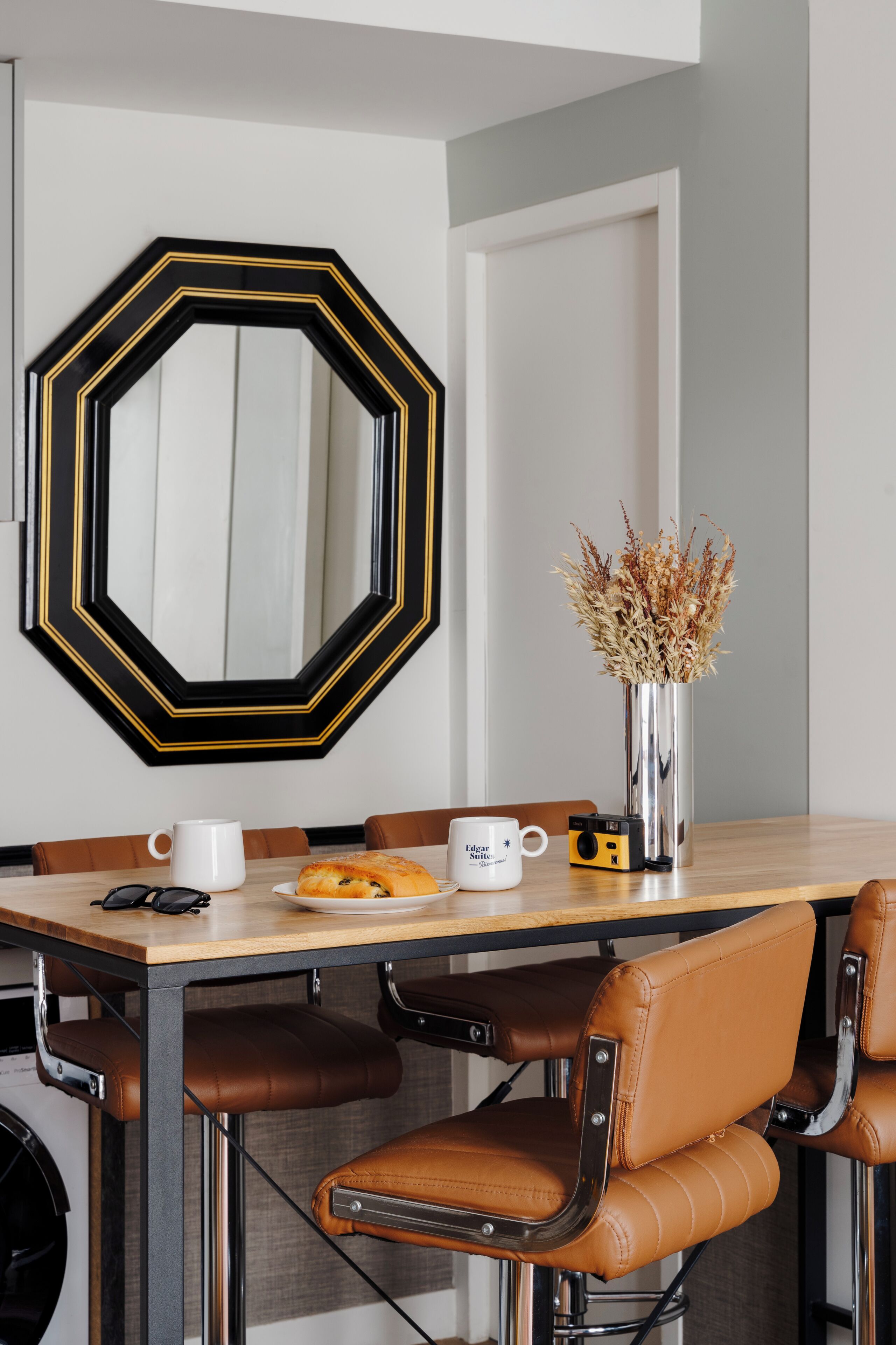 A dining area with a wooden table, leather chairs, a vase with dried plants, and a camera on the table.