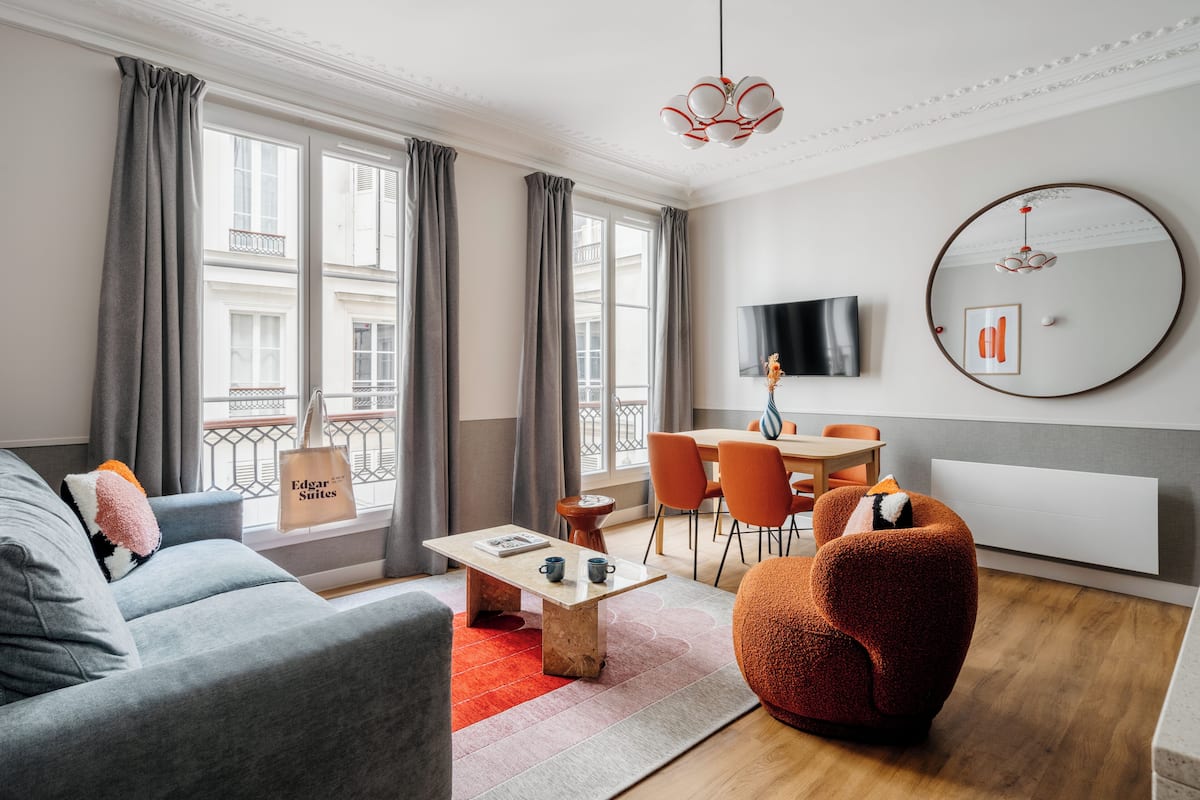 A modern living room with a grey sofa, a red armchair, a wooden coffee table, and a dining area with orange chairs. A large round mirror and a flat-screen TV are mounted on the wall.