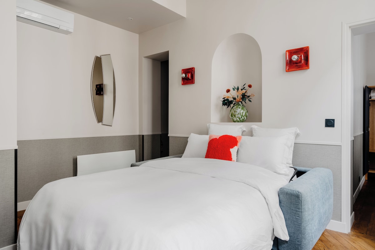 Habitación de hotel con una cama grande, un espejo, un jarrón con flores y un interruptor de luz rojo.