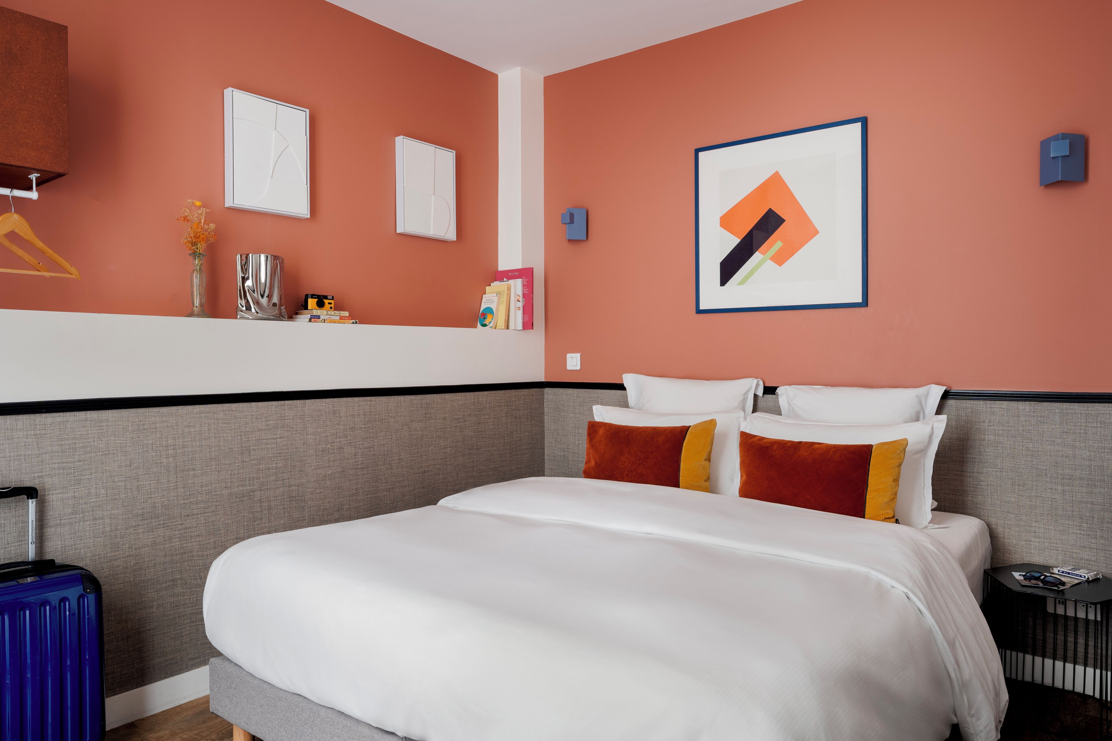 Hotellihuone, jossa on suuri sänky, yöpöytä, matkalaukku ja seinä, jossa on kehystettyjä taideteoksia.