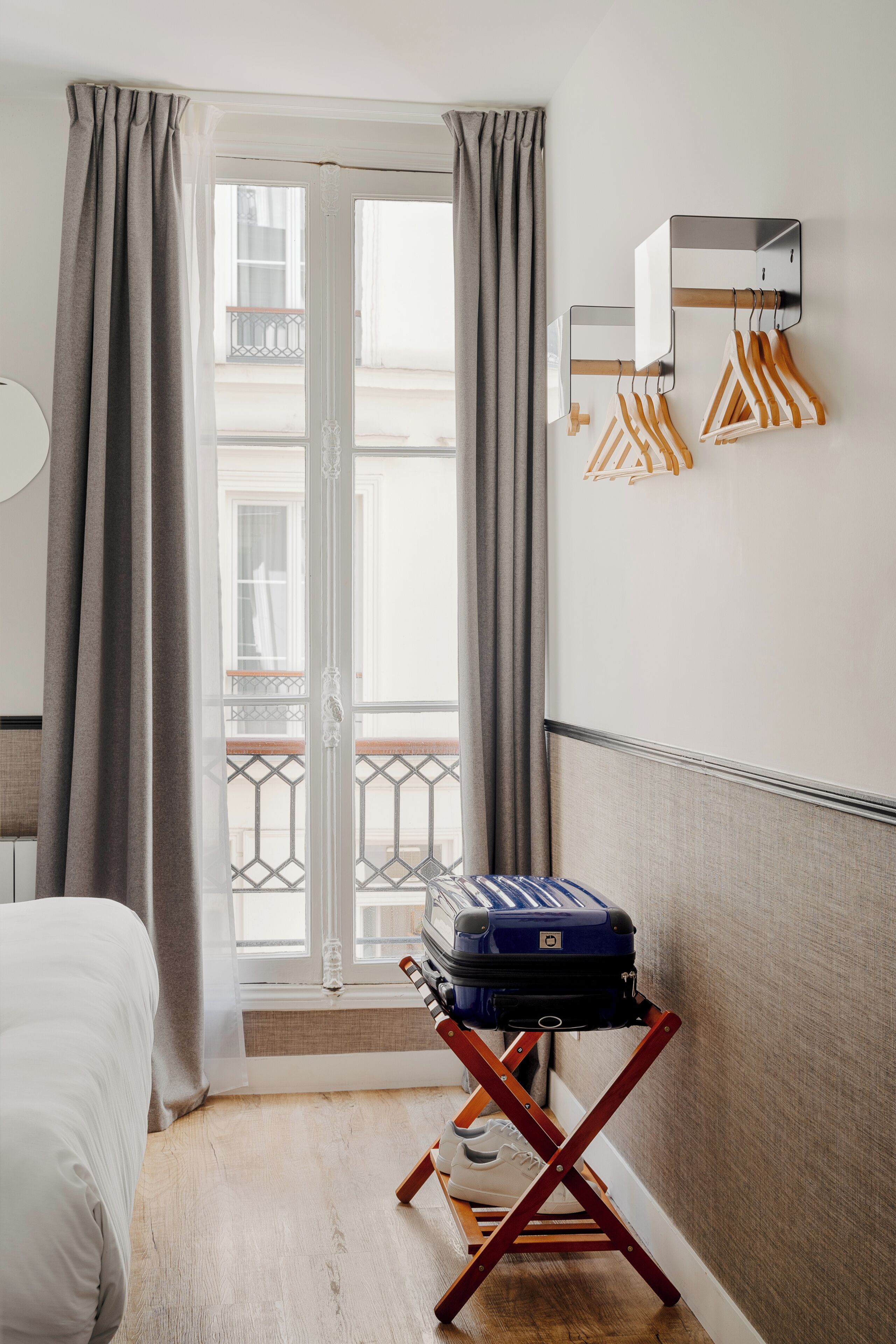 A hotel room with a bed, a suitcase on a wooden stand, a wall-mounted coat rack, and a view through a door with curtains.