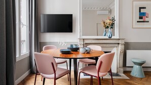 Un área de comedor con una mesa redonda, cuatro sillas, un televisor de pantalla plana montado en la pared, una chimenea y un cuadro abstracto enmarcado.