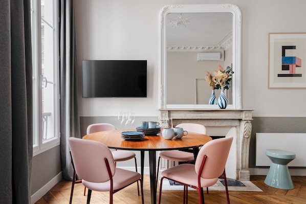 A dining area with a round wooden table, pink chairs, a flat-screen TV, a vase with flowers, and a fireplace.