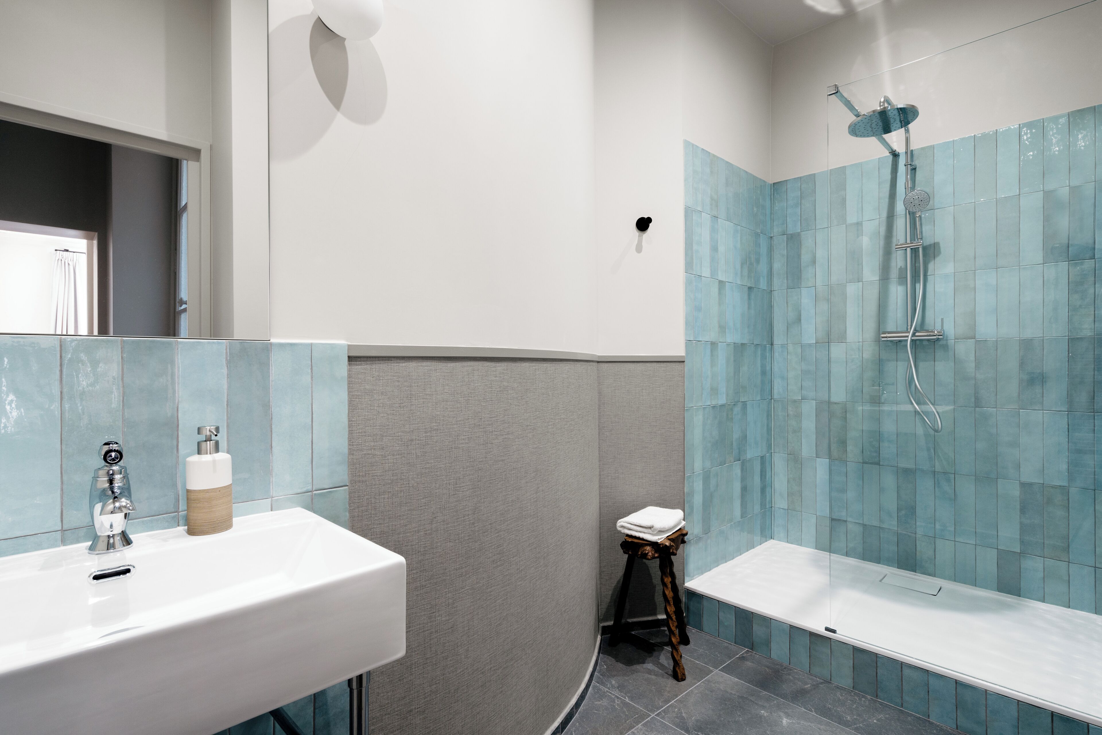 Ein Badezimmer mit einem weißen Waschbecken, einem Spiegel, einem Handtuchhalter, einer Dusche mit Glastrennwand und einem kleinen Hocker.