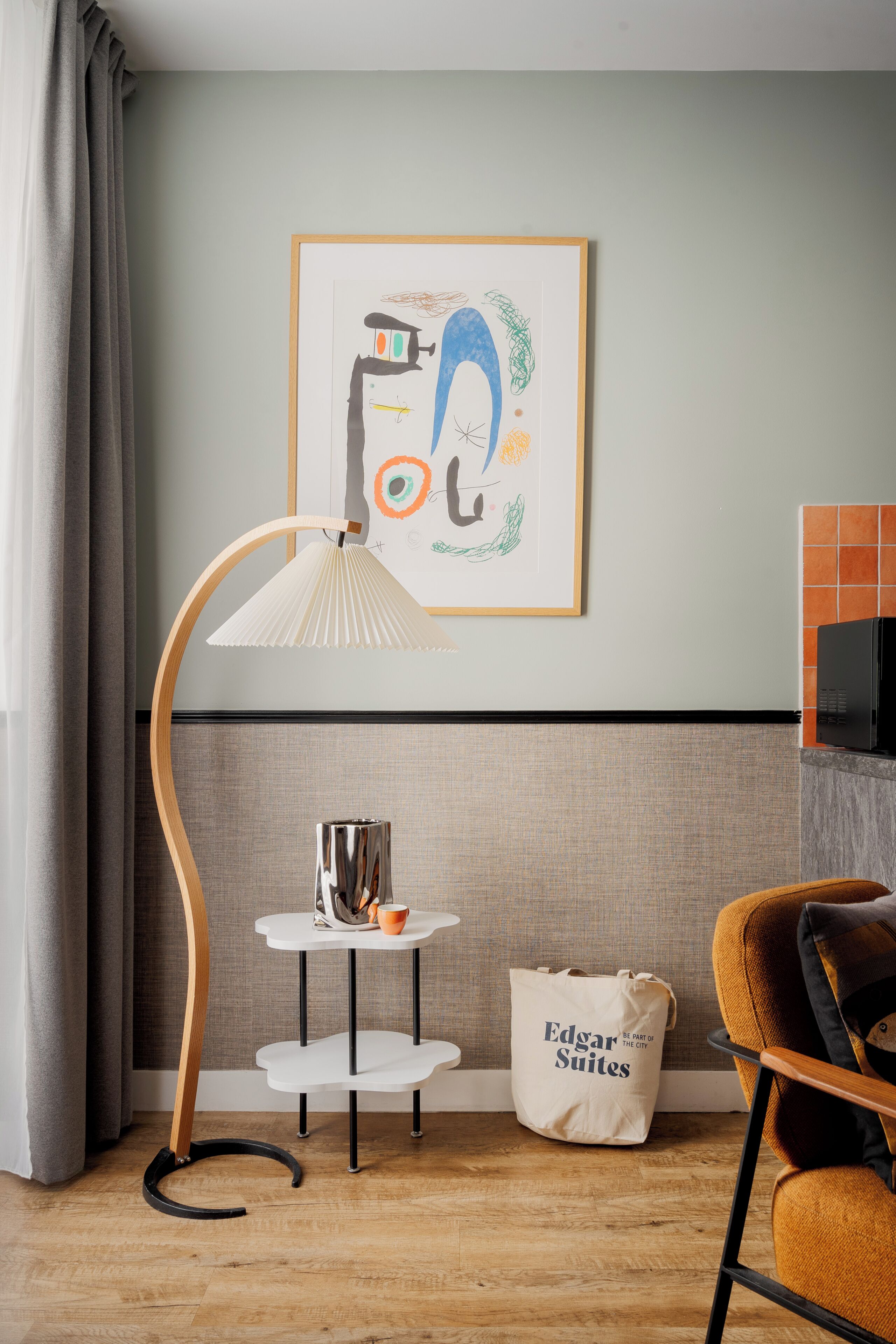 A modern living room with a grey sofa, a wooden floor lamp, a small white table with a cup, and a framed abstract artwork on the wall.
