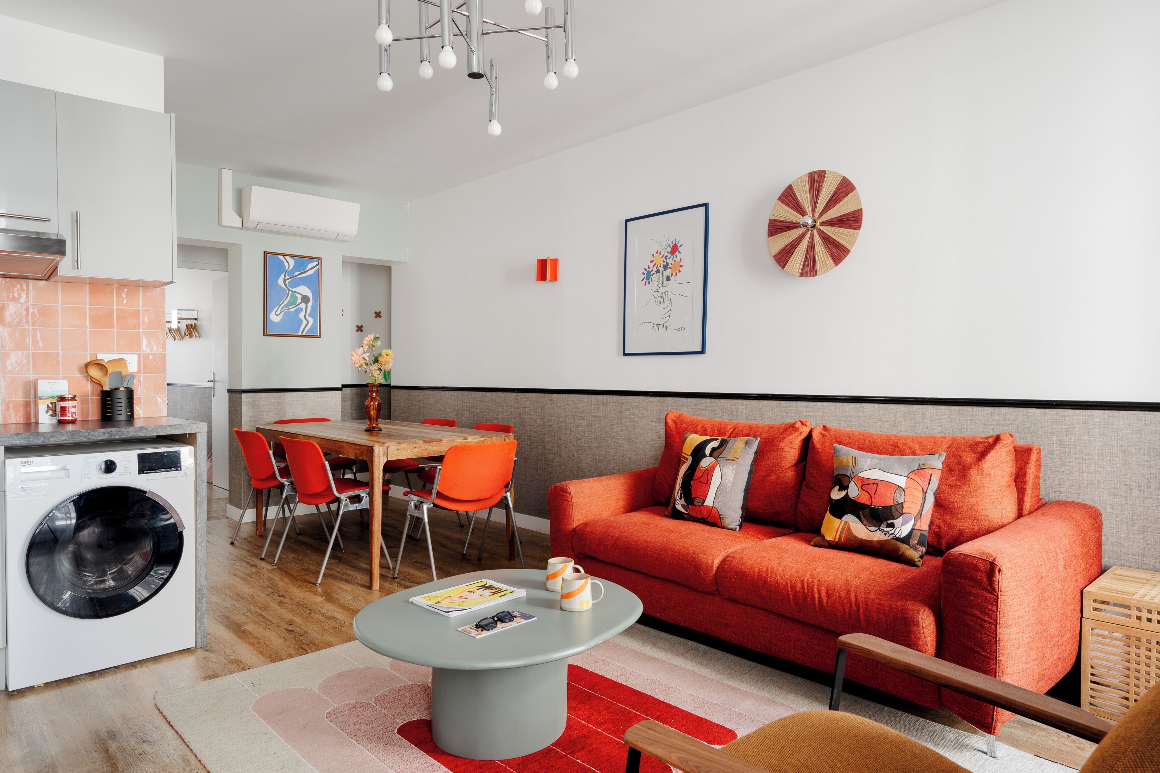 Un salon moderne avec un canapé rouge, un coin repas avec des chaises orange et une cuisine équipée d’un lave-linge.