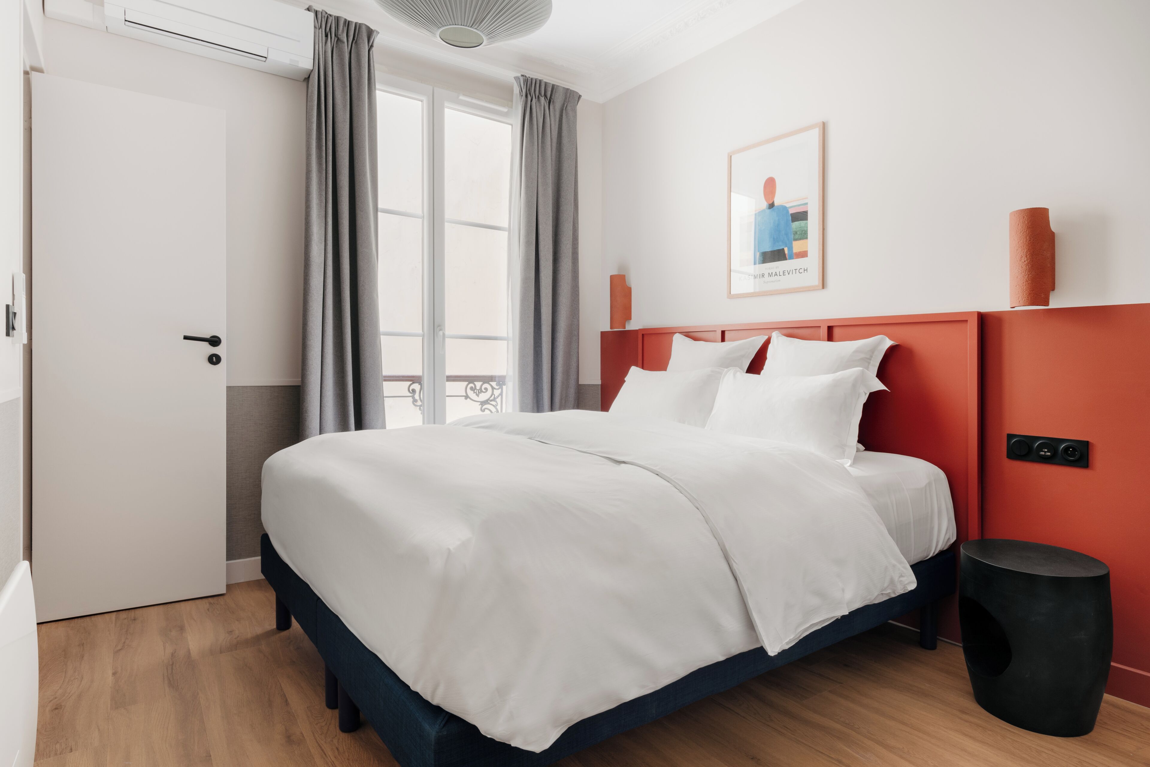 Una camera d'albergo con un letto, una porta, una finestra con tende, un'opera d'arte appesa al muro e un comodino.