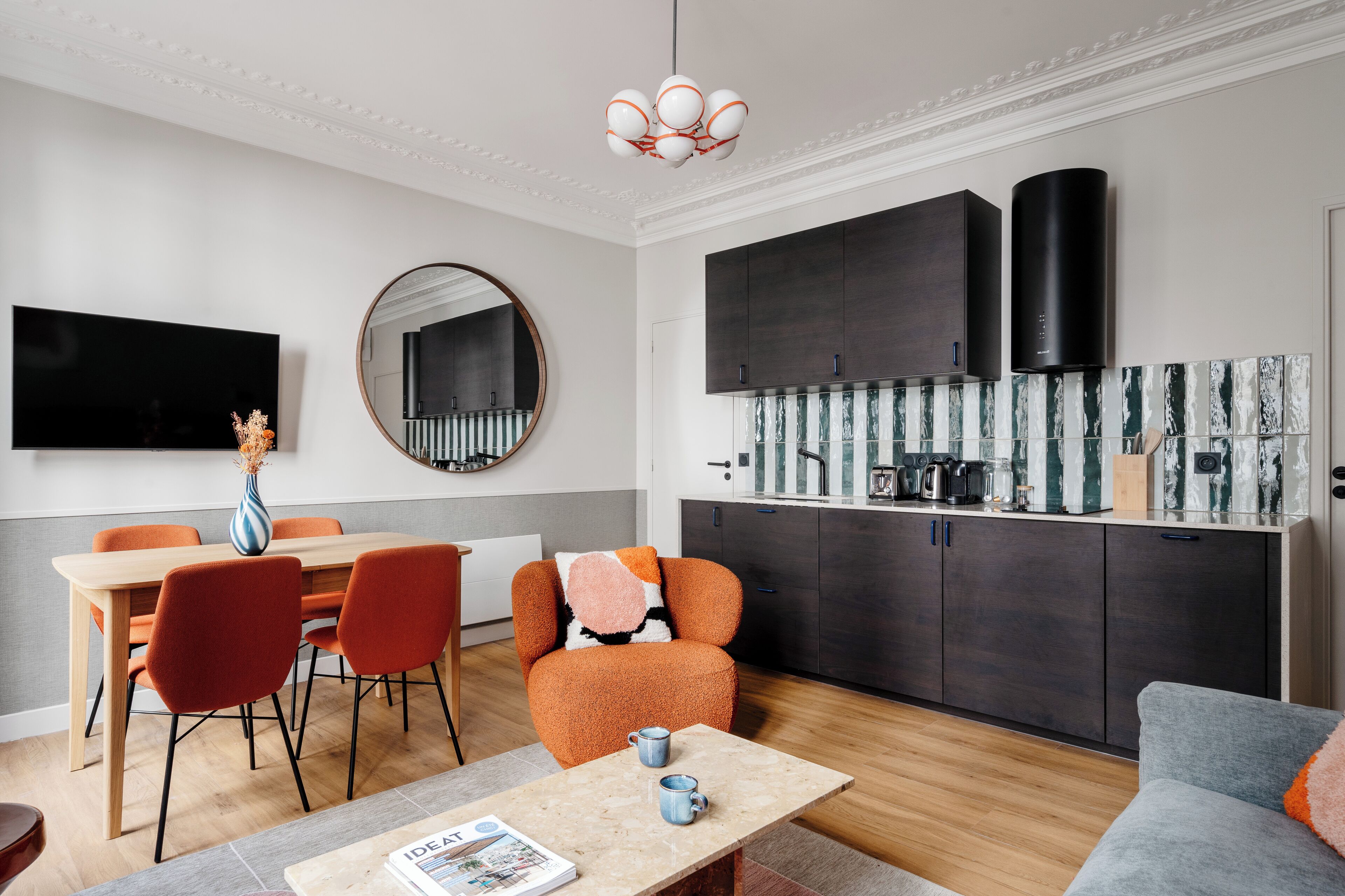 A modern living room with a dining area, a kitchen with dark cabinets, and a large flat-screen TV mounted on the wall.