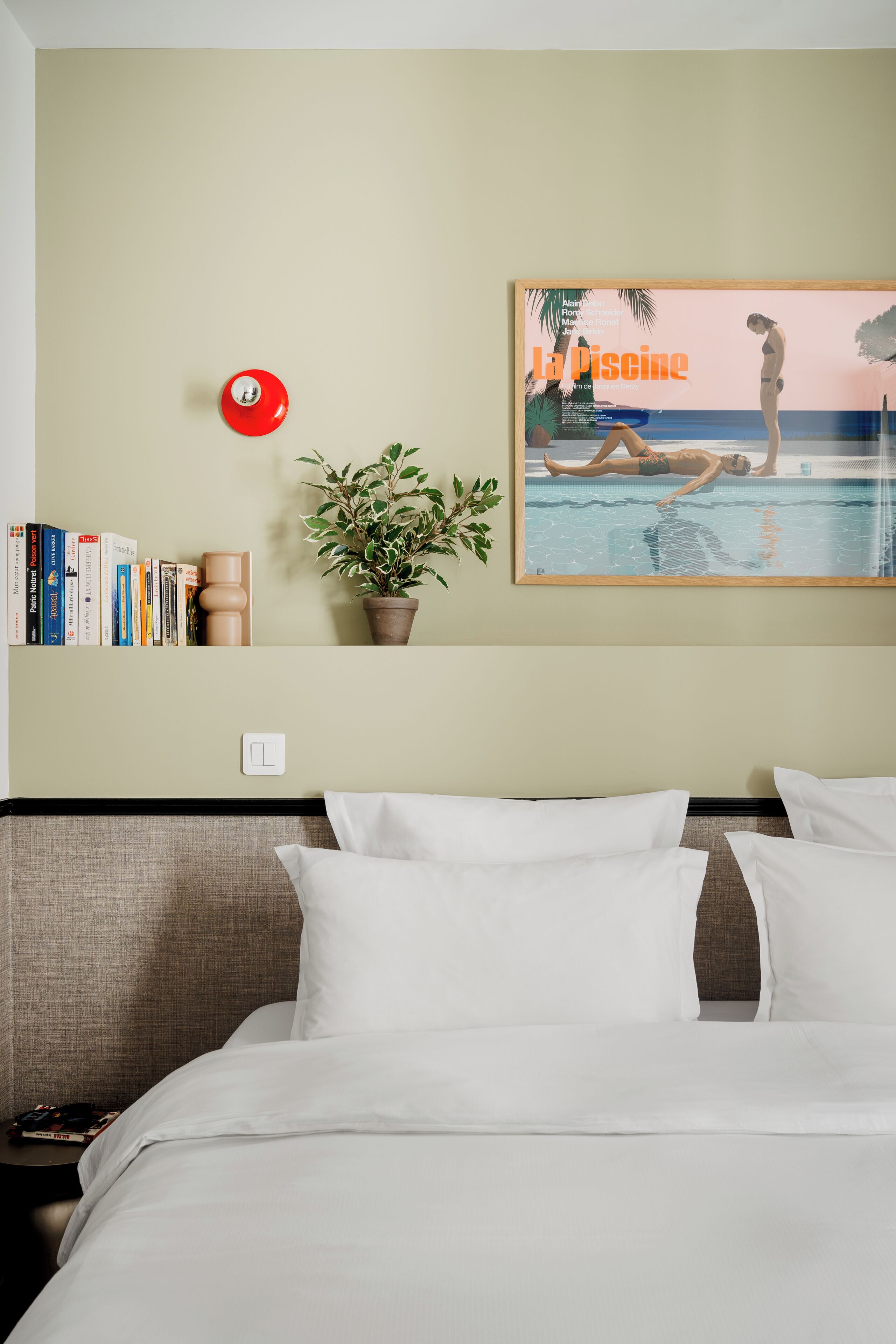 Una habitación de hotel con una cama, una mesita de noche, una lámpara de pared, un cuadro enmarcado, una planta en maceta y una estantería.