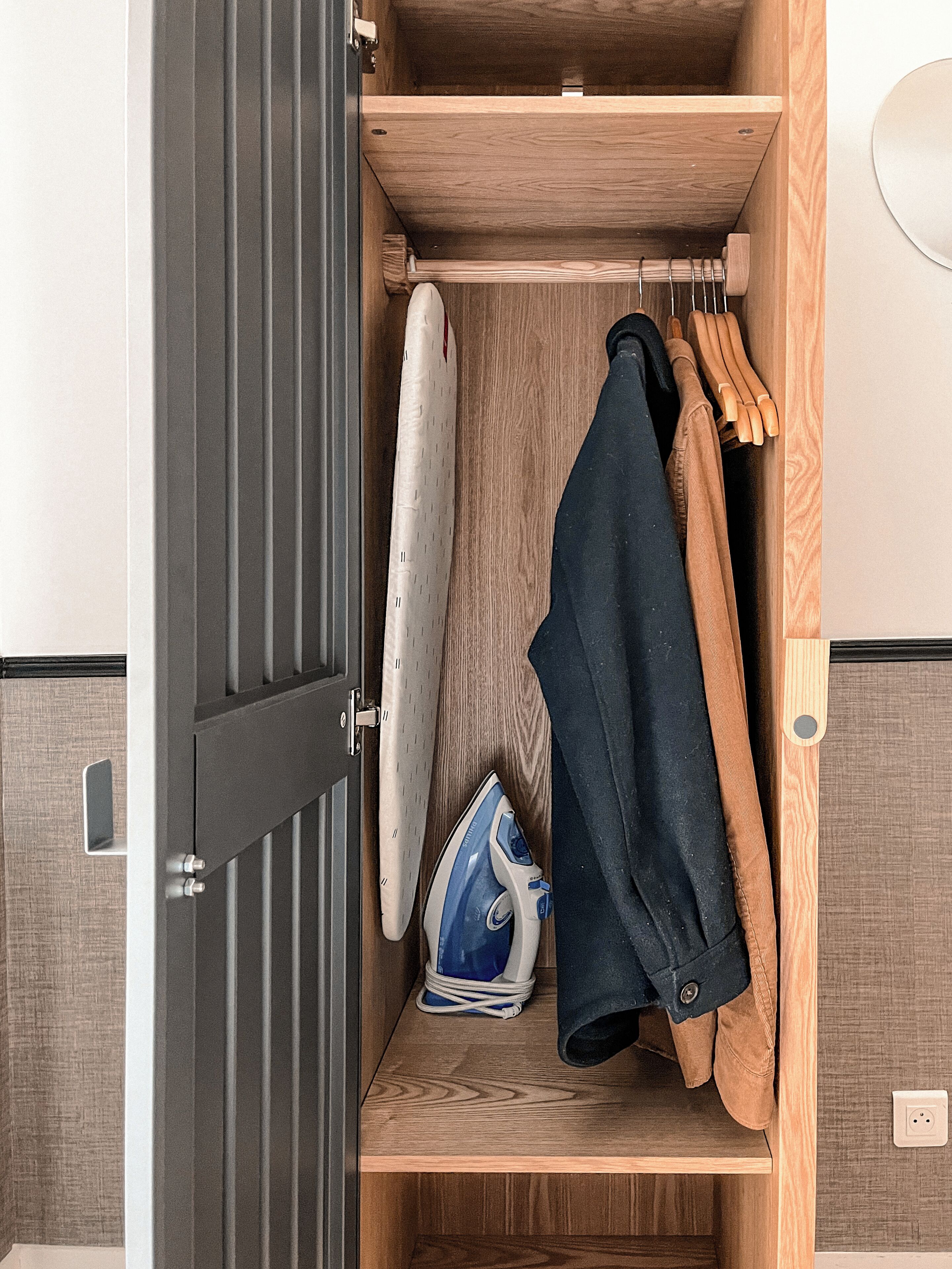 A wooden wardrobe with a hanging rack, an ironing board, and a pair of shoes.
