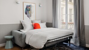 Un dormitorio con cama, almohadas, una mesita de noche, una obra de arte enmarcada y una ventana con cortinas.