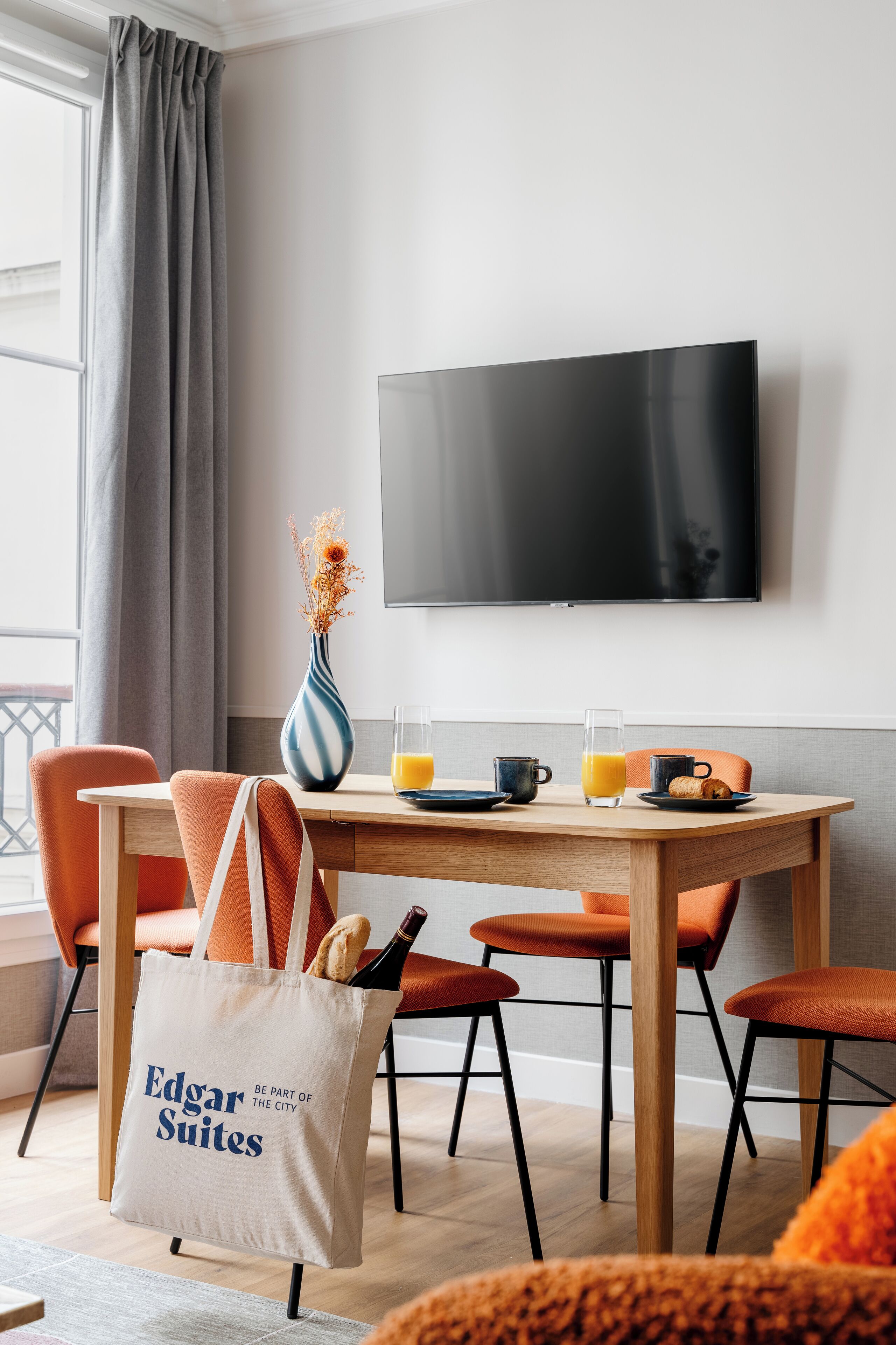 A modern dining area with a wooden table, orange chairs, a vase with dried flowers, a glass of orange juice, a bottle of wine, and a tote bag with "Edgar Suites" text.