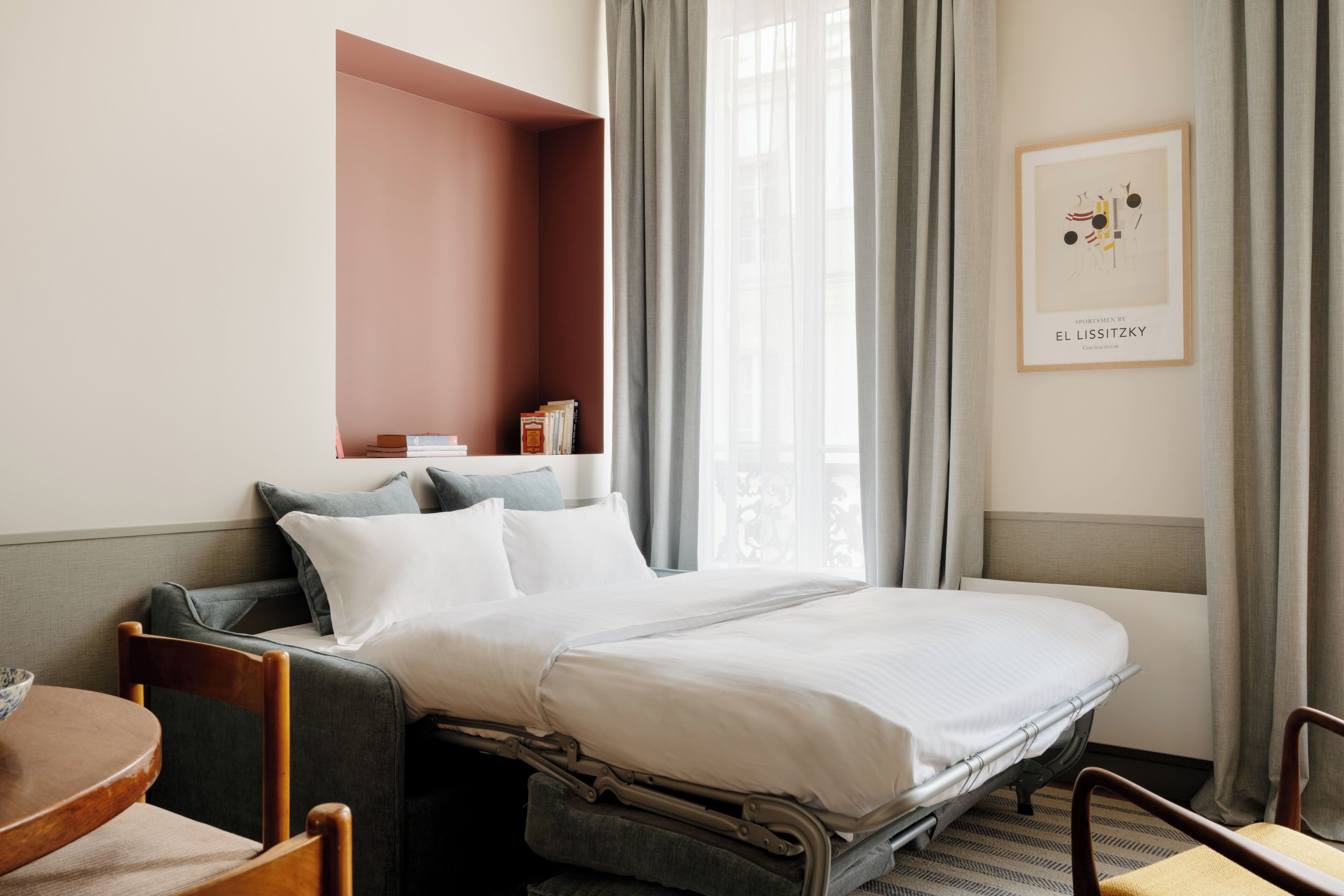 A hotel room with a bed, a seating area with chairs, a small table, a bookshelf, and a window with curtains.