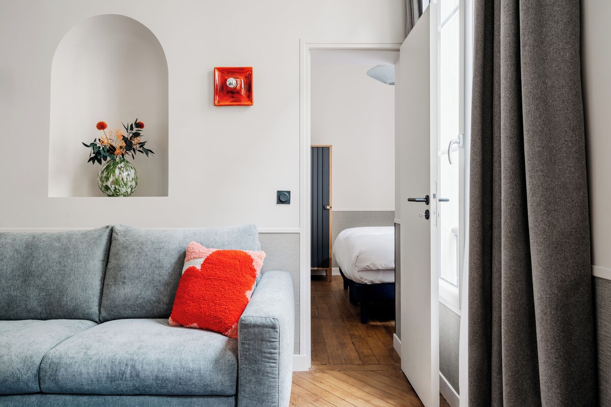 Un salon moderne avec un canapé gris, un coussin rouge, un vase de fleurs et une porte donnant sur une chambre.