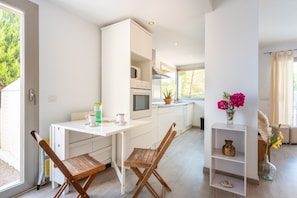 Une cuisine moderne avec des meubles blancs, un coin repas avec des chaises en bois et une petite étagère ornée d’objets décoratifs.