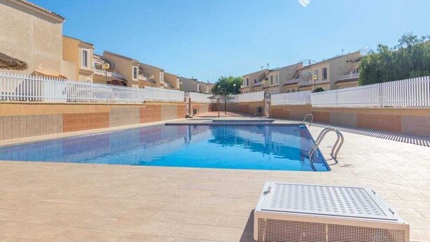 A swimming pool with a tiled deck, surrounded by buildings with balconies and a white fence.
