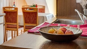 Huoneessa on ikkuna ja ovi, ja siellä on puinen pöytä, jonka päällä on kulho täynnä hedelmiä, vaaleanpunainen liina ja puiset tuolit.