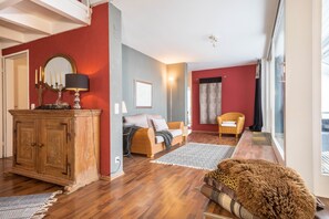 Ein Zimmer mit einem Holzkasten, einem Sofa, einem Sessel, einem Teppich und einem großen Fenster.