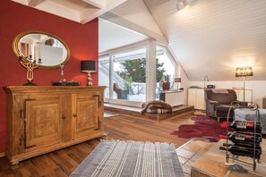 Ein Zimmer mit einem Holzkabinett, einem Spiegel, einer Lampe, einem Weinkühler, einem Sessel und einem großen Fenster.