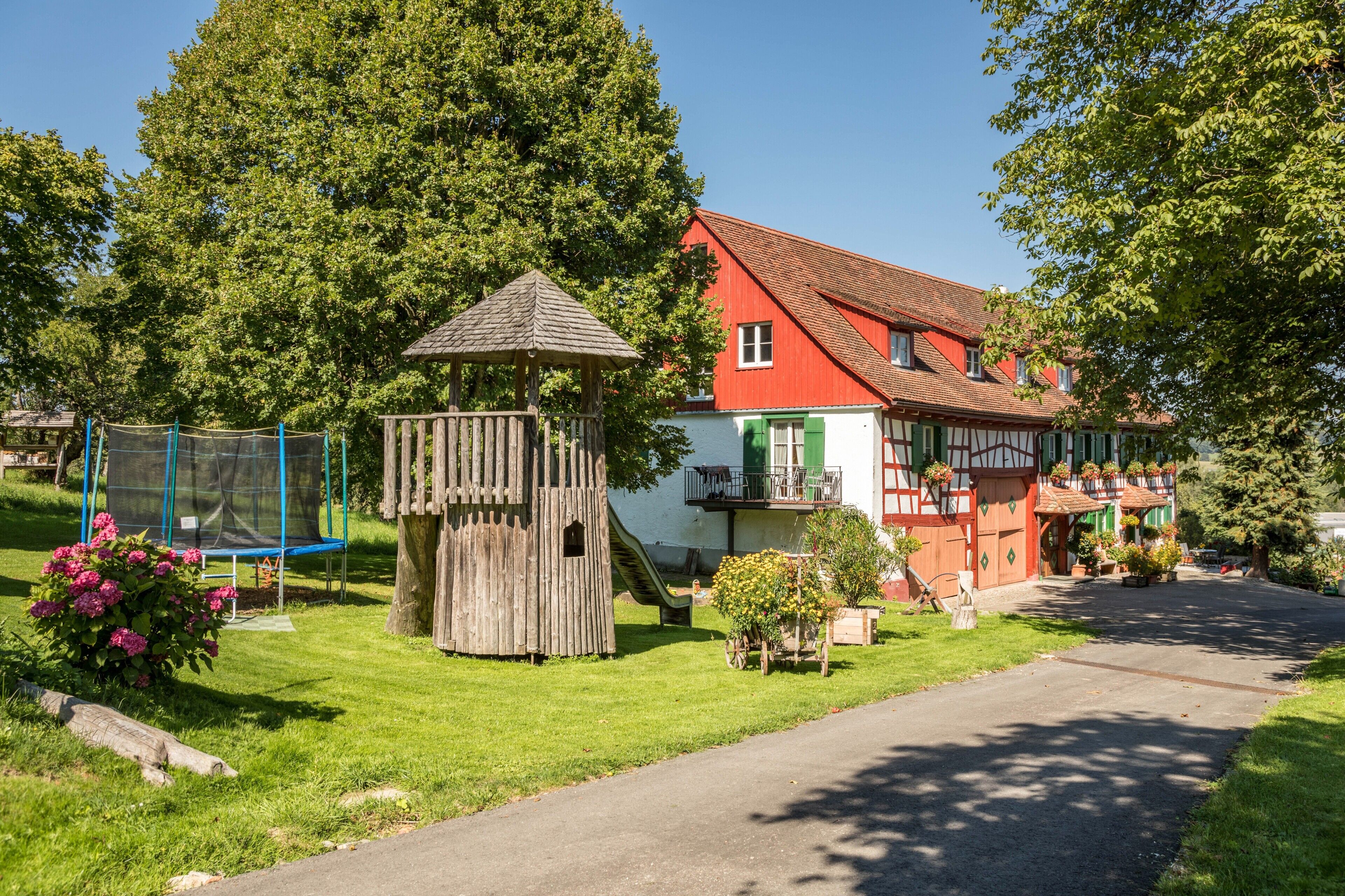 Ein traditionelles Haus mit rotem Dach, einer hölzernen Spielanlage und einem Garten mit Blumen.