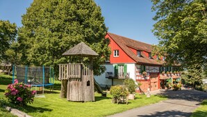 A traditional house with a red roof, a wooden play structure, and a garden with flowers. - Holiday Apartment "Risthof 1" with Wi-Fi, Shared Garden, Sauna & Playground (Owingen)