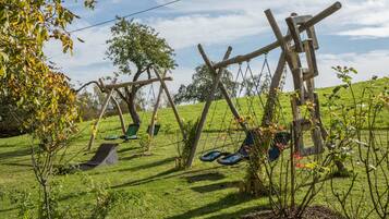 Ein grünes Areal im Freien mit Holzschaukeln und einer kleinen Rutsche.