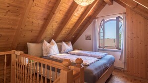 A wooden attic bedroom with a sloped ceiling, a bed with a headboard, a window with curtains, and a wooden railing. - Holiday Apartment ‘Haus Desor’ with Lake View, Wi-Fi, Balcony (Kressbronn)