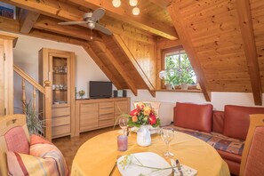 Une pièce chaleureuse avec une table à manger dressée pour deux personnes, un buffet en bois, un ventilateur de plafond et une fenêtre donnant sur un paysage verdoyant.