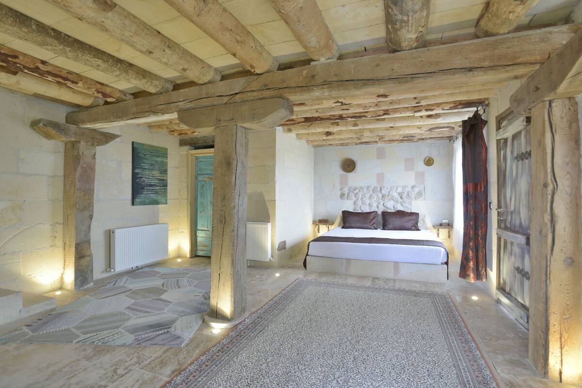 A rustic bedroom with exposed wooden beams, a stone wall, a bed with a white duvet, and a patterned rug.