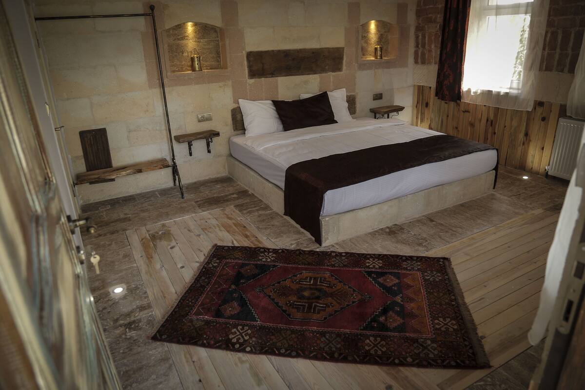 A bedroom with a large bed, a wooden floor, a patterned rug, and a brick wall.