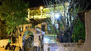 Um arco de pedra que dá acesso a um edifício com uma escadaria de madeira e uma árvore em primeiro plano.