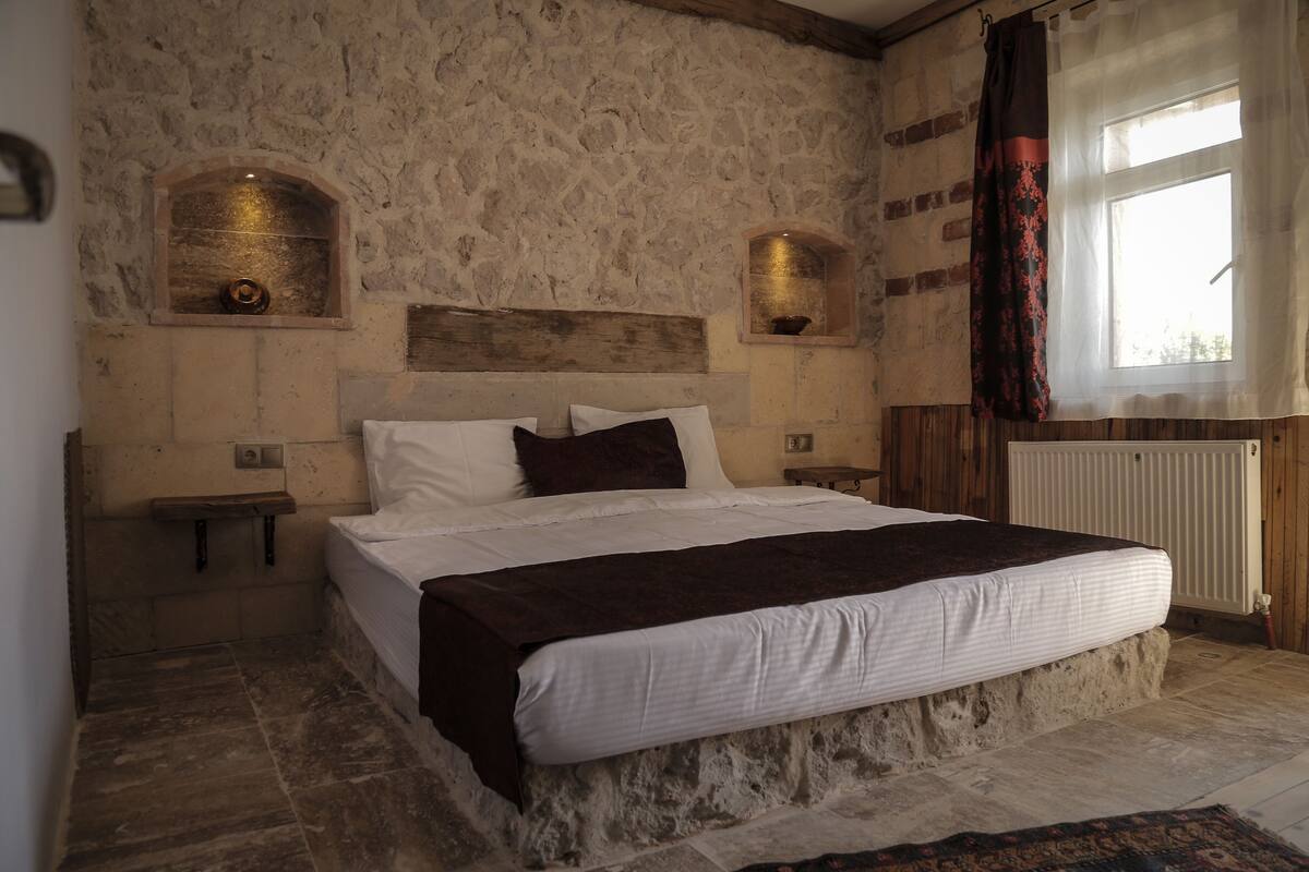 A bedroom with a stone wall, a wooden headboard, a large bed with white and brown bedding, two bedside tables, and a window with curtains.