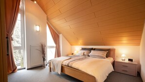 A bedroom with a wooden ceiling, a large bed with multiple pillows, bedside tables with lamps, and a window with curtains.