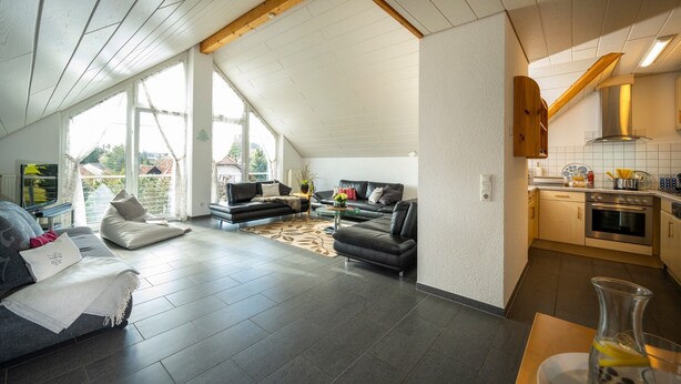 A spacious living area with a high ceiling, a kitchen with modern appliances, and a large window providing natural light.