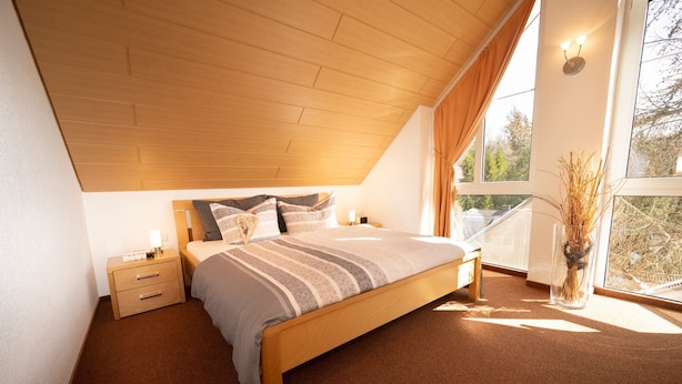 Ein Schlafzimmer mit Holzboden, einem Bett mit gestreifter Bettwäsche, einem Nachttisch, einer Nachttischlampe und einem Fenster mit Blick auf Bäume.