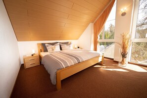 A bedroom with a wooden ceiling, a bed with striped bedding, a nightstand, a bedside lamp, and a window with a view of trees.