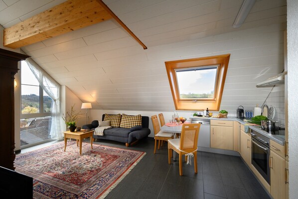 A cozy loft-style room with a dining area, a sofa, and a kitchen with a large window.