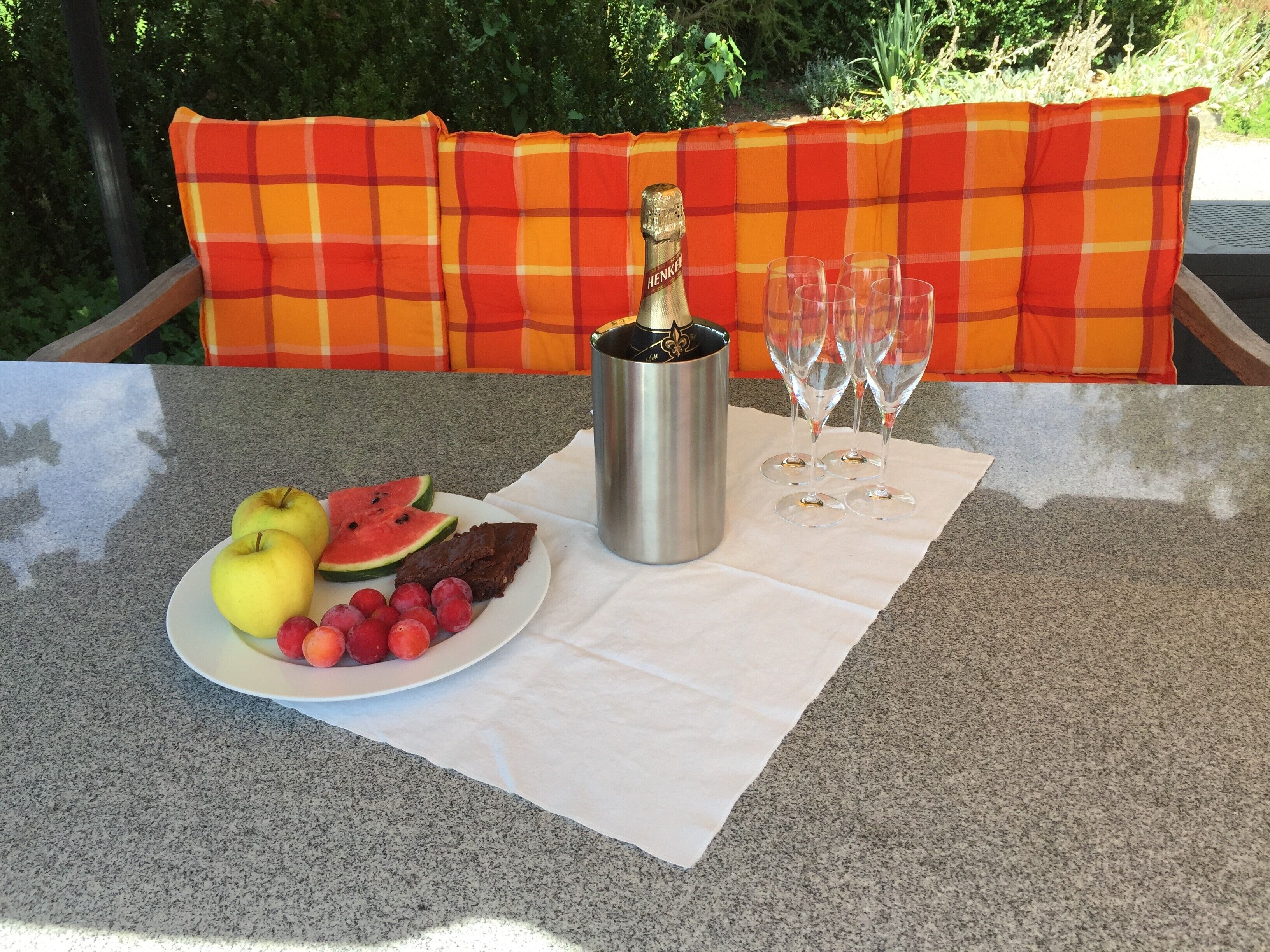 Una mesa con una botella de champán, cuatro copas, un plato con frutas y un postre de chocolate, y un cojín a cuadros naranja y rojo al fondo.