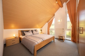 Een slaapkamer met een schuine kap, een houten bed, een nachtkastje, een klein krukje en uitzicht op het buitengebied door een raam.