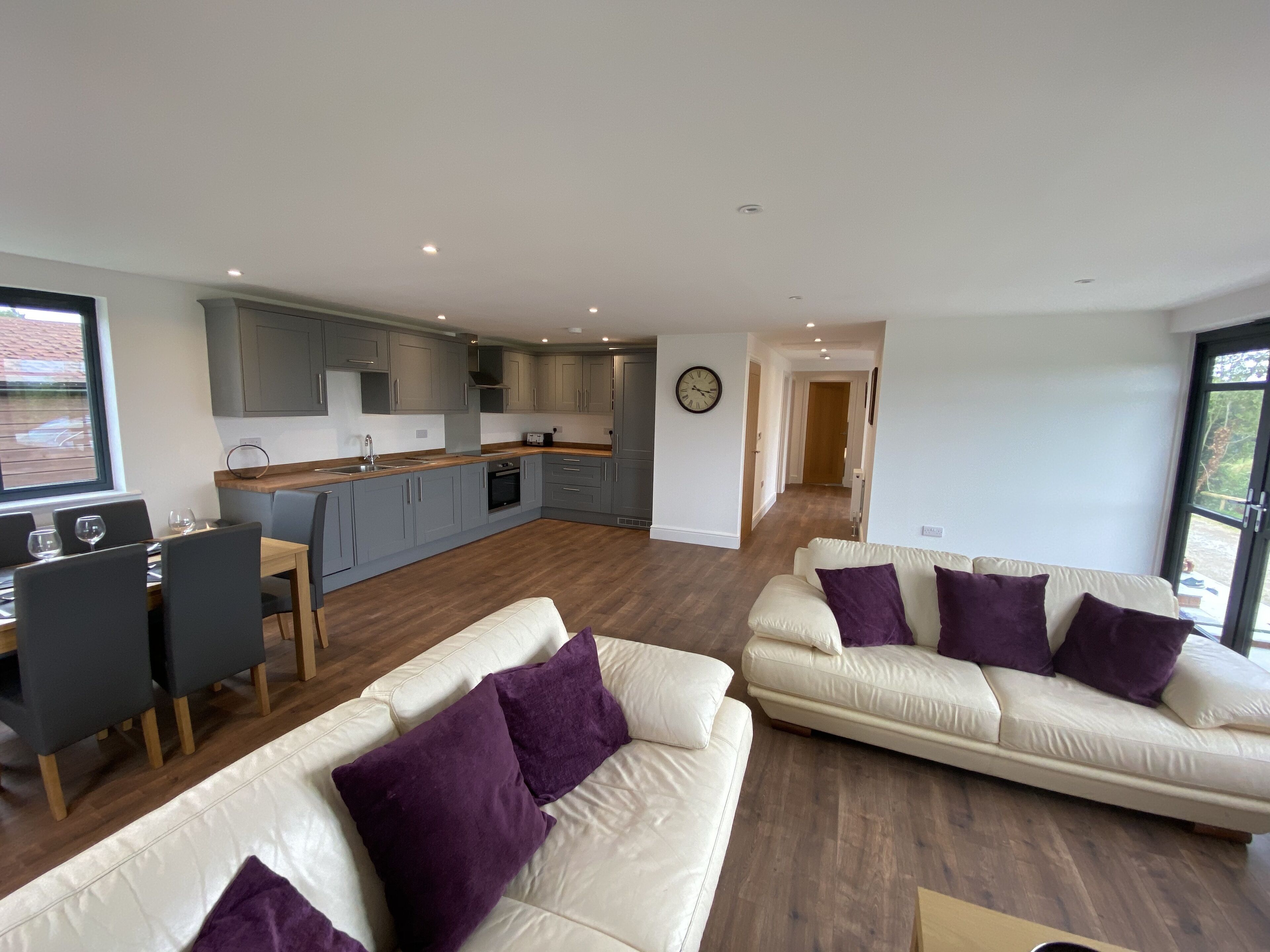 A modern living room with a sofa and chairs, a dining area with a table and chairs, and a kitchen with grey cabinets and a window.
