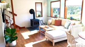 A cozy living room with a sofa, ottoman, and a small table. There is a fireplace and a window with a view of greenery. - The Shell Shack-Darling and peaceful cottage (Pacific City)