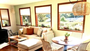 A living room with a sofa, a dining table with chairs, and large windows with a view of greenery. - The Shell Shack-Darling and peaceful cottage (Pacific City)